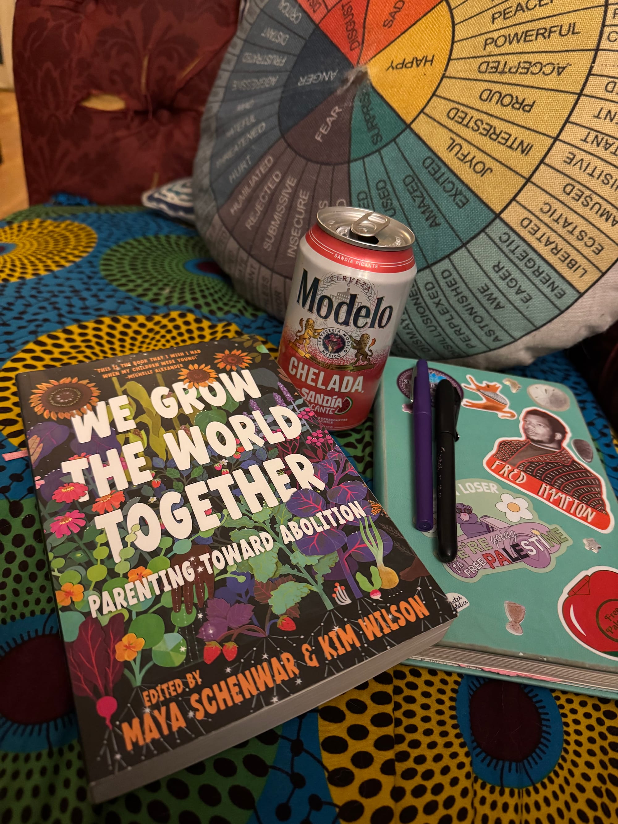 a copy of the book ‘we grow the world together: parenting towards abolition’ (edited by Maya Shenwar and Kim Wilson with beautiful cover art by Olly Costello) sits on a surface of brightly colored African fabric, next to a can of Modelo  Sandia Chelada, with a colorful circular feelings pillow in the background.