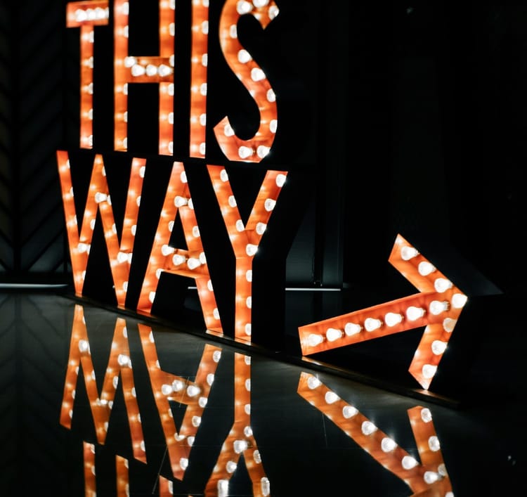 ‘This way’ sign lit with bulbs in a dark space w/ an arrow pointing up towards the right; sign reflected onp surface below.